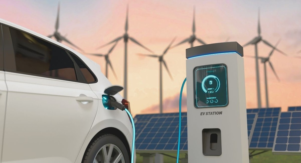 Voiture électrique branchée à une borne de recharge devant des éoliennes et des panneaux solaires